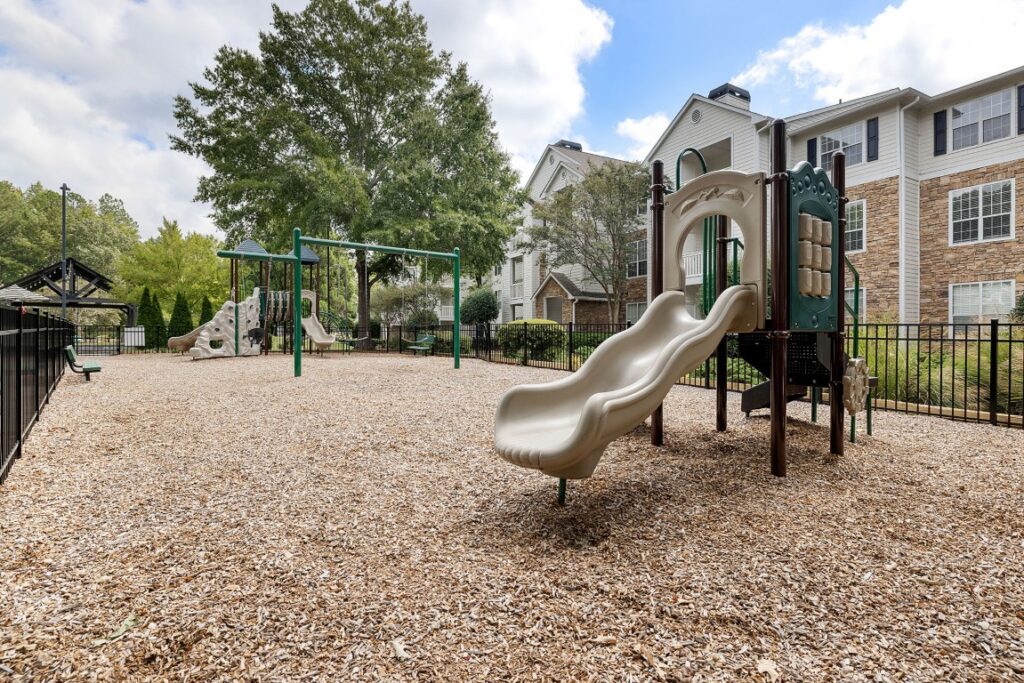 Playground with wood chip ground cover featuring slides, climbing structures, and swing sets in front of an apartment building.