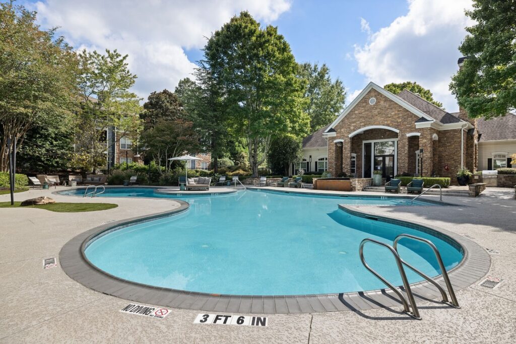 A large, kidney-shaped outdoor swimming pool surrounded by a concrete deck, patio furniture, and a stone clubhouse building.