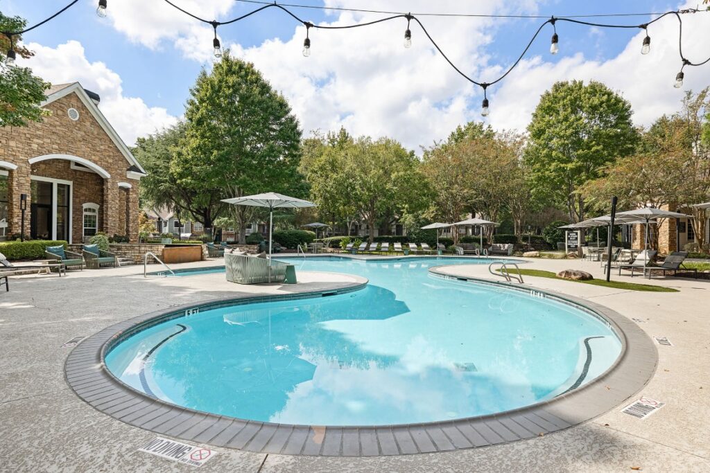 Curved swimming pool on a patio with lounge chairs, umbrellas, and string lights overhead.