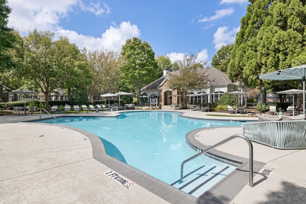 A curved swimming pool with a stone patio, lounge chairs, and a clubhouse building in the background.