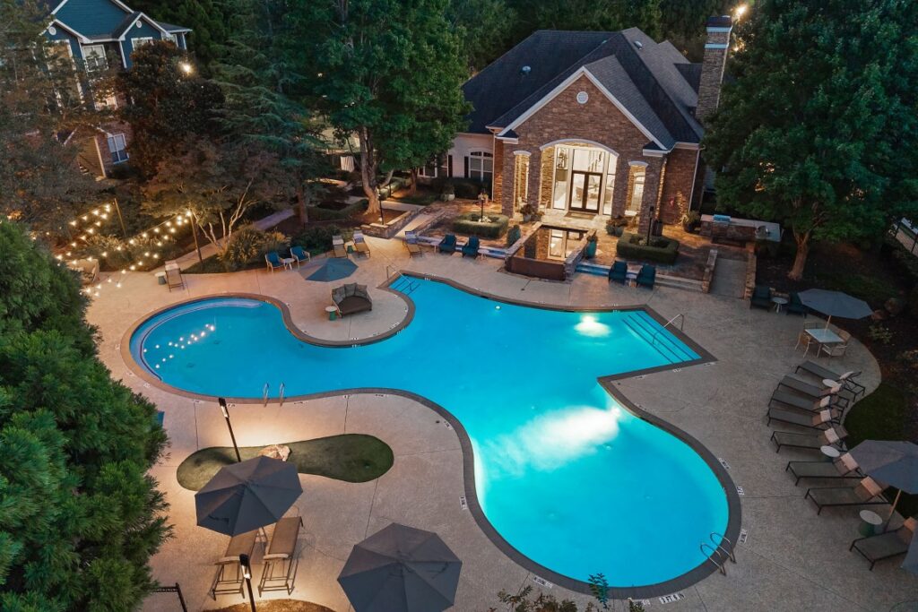 Aerial view of a brightly lit swimming pool and surrounding patio at dusk, with lounge chairs, umbrellas, and a clubhouse building in the background.