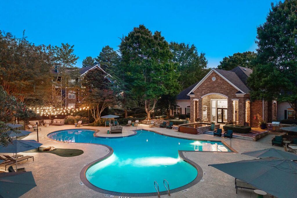A resort-style swimming pool at dusk, surrounded by stone patio, lounge chairs, umbrellas, and the clubhouse building.