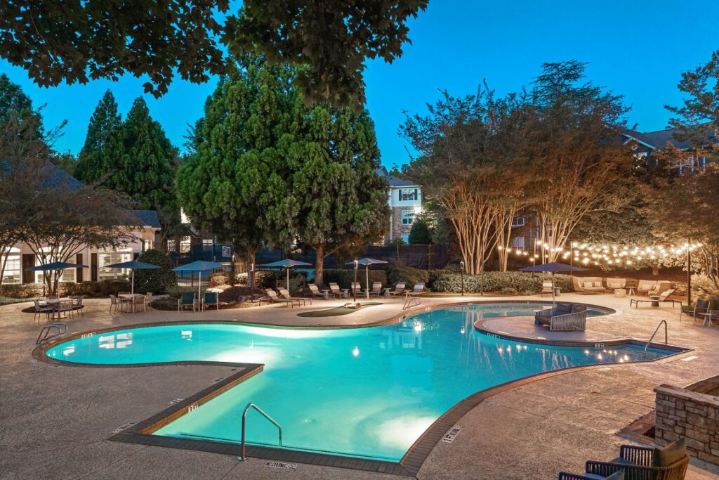 A curved outdoor swimming pool at twilight, surrounded by lounge chairs, patio furniture, and string lights.