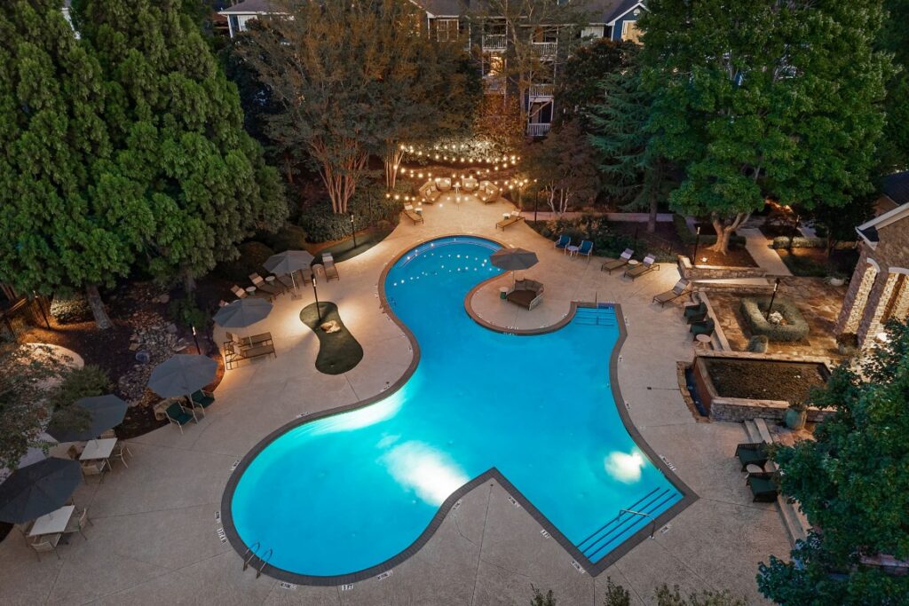 Aerial view of an illuminated, freeform swimming pool surrounded by a patio, trees, and string lights at night.