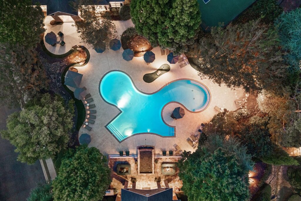 Aerial view of a kidney-shaped swimming pool at night surrounded by trees, lounge chairs, and umbrellas.