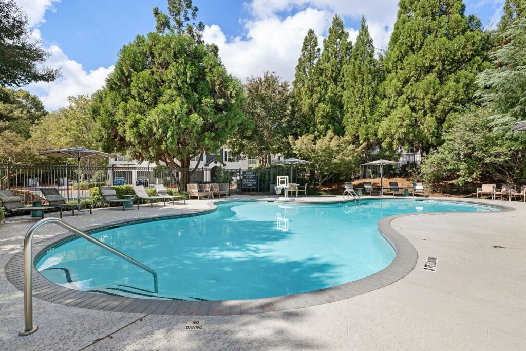 A kidney-shaped outdoor swimming pool surrounded by lounge chairs, patio tables, and trees.