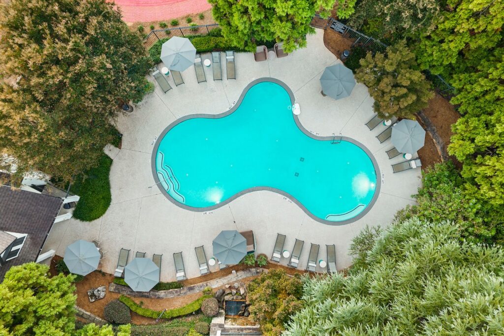 An aerial view of a clover-shaped swimming pool surrounded by a concrete deck with lounge chairs and patio umbrellas, all nestled within a lush, wooded area.