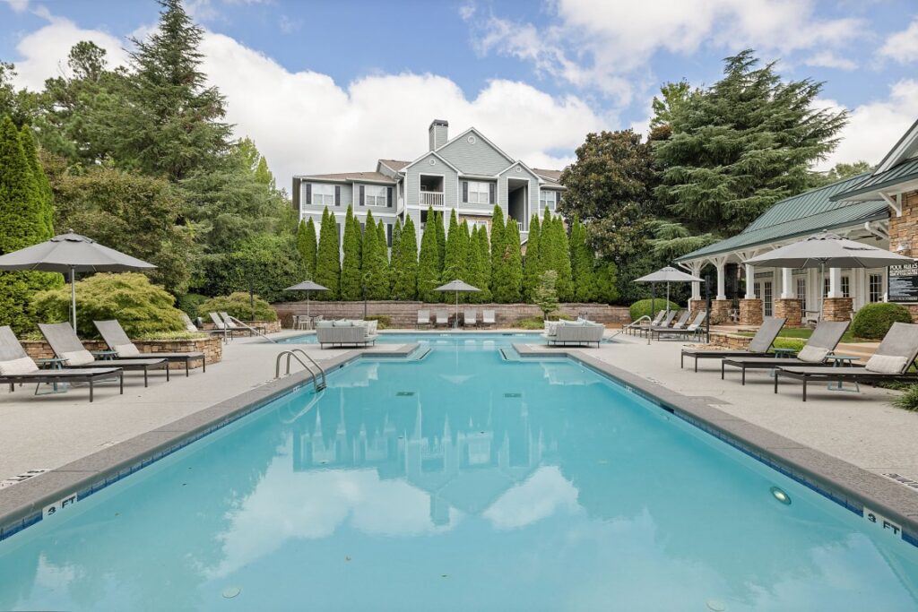 A rectangular swimming pool surrounded by a patio with lounge chairs, umbrellas, and a tall apartment building in the background.