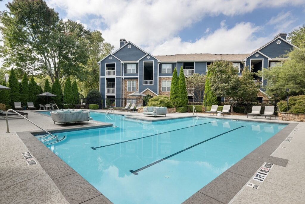 A blue apartment building stands behind a large swimming pool with surrounding patio furniture and lounge chairs.