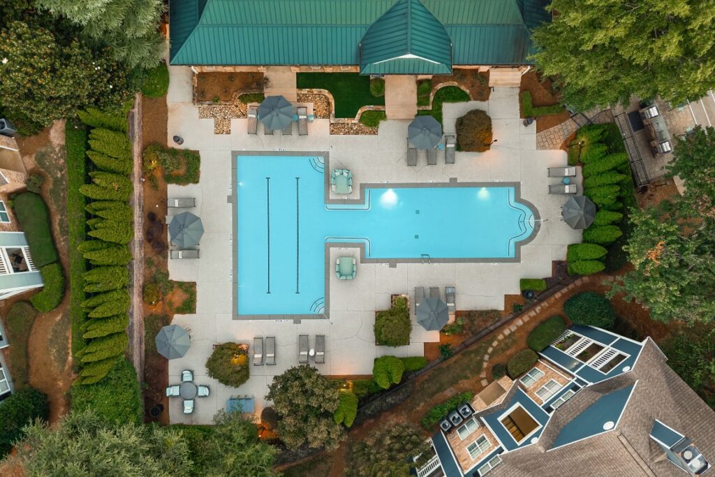 Aerial view of a T-shaped swimming pool surrounded by a patio with lounge chairs, umbrellas, and dining tables, situated next to two multi-story buildings and trees.