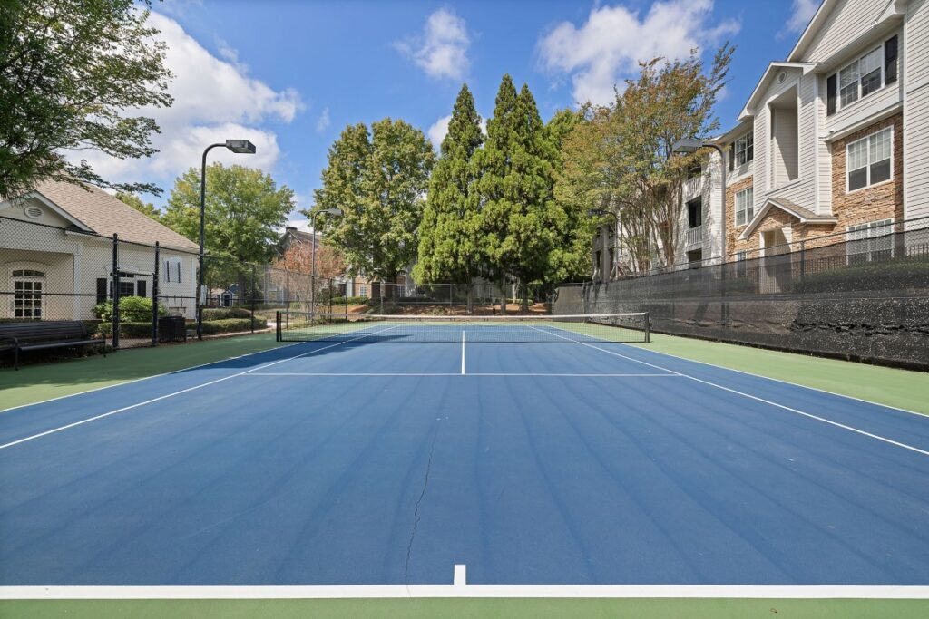 A blue and green tennis court surrounded by a black chain-link fence, with apartment buildings and trees in the background.