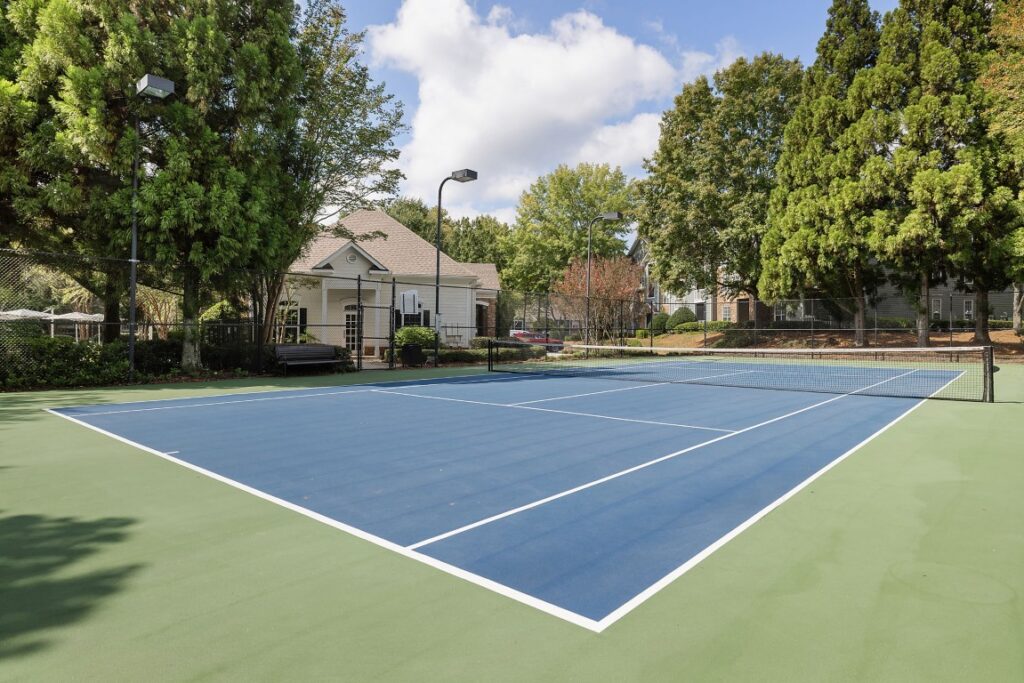 A blue and green outdoor tennis court surrounded by tall trees and a small clubhouse.