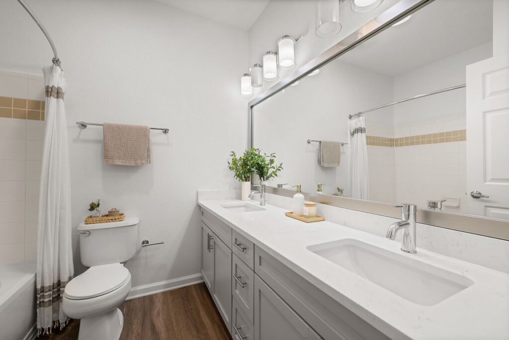Bathroom with a double-sink white vanity, gray cabinets, a large mirror, and a toilet next to a shower with a white curtain.