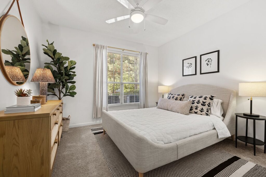 A bedroom with a light-colored upholstered bed, a wooden dresser, a large round mirror, and a window with white curtains.