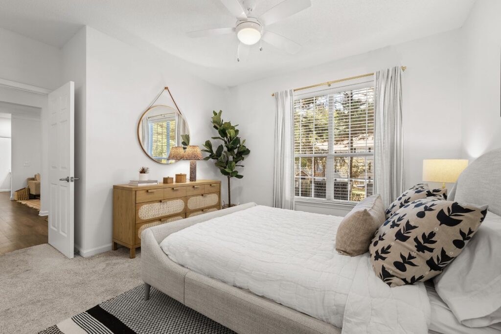 A bedroom featuring a light-colored upholstered bed, a wooden dresser with a circular mirror, and a window with white curtains.