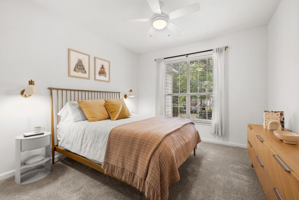 A bedroom with a brass bed frame, gold-toned pillows, a textured throw blanket, a white nightstand, and a wooden dresser under a ceiling fan.