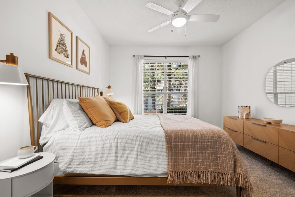 A bedroom with a queen bed featuring a brass headboard, gold-toned pillows, and a textured throw blanket, flanked by a white nightstand and a wooden dresser with a round mirror.