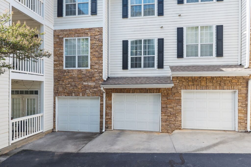 A three-story building exterior with white siding, stone accents, three white garage doors, and a covered balcony.