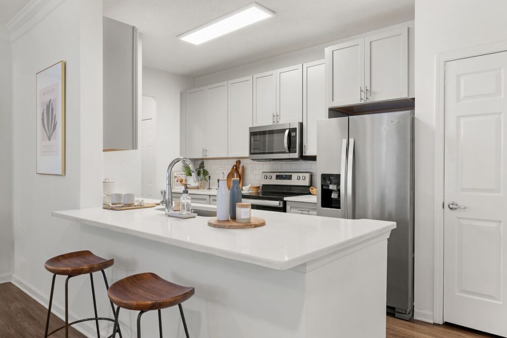 A modern kitchen with a white quartz breakfast bar, two wooden stools, stainless steel appliances, and light gray cabinets.