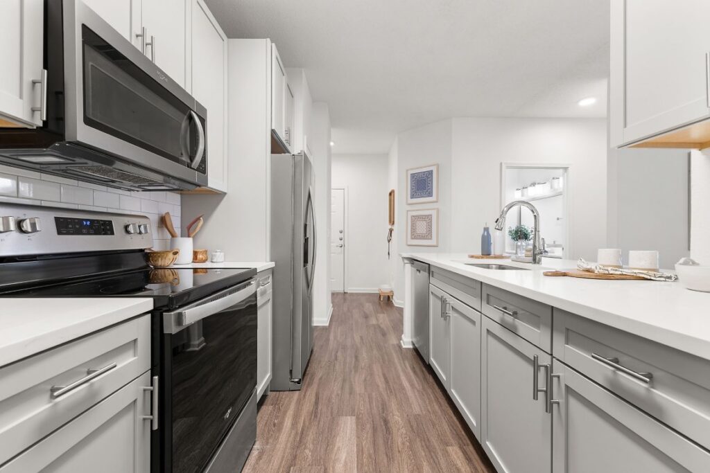 A narrow galley kitchen with white cabinets, stainless steel appliances, and wood-look flooring.