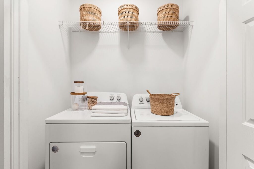 A laundry closet with a white washer and dryer, topped with folded towels, small jars, and a woven basket, with three identical baskets on a wire shelf above.