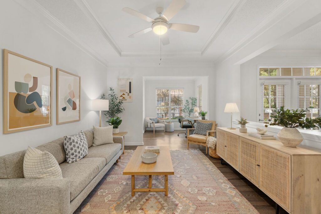 Living room with a light gray sofa, wooden coffee table, patterned area rug, and a long wooden console table, opening into a small office space with a desk and chair.
