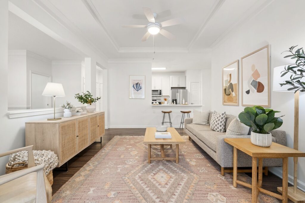 A bright living room with a beige sofa, a low wooden coffee table, and a cane-front sideboard, opening into a kitchen with a breakfast bar.