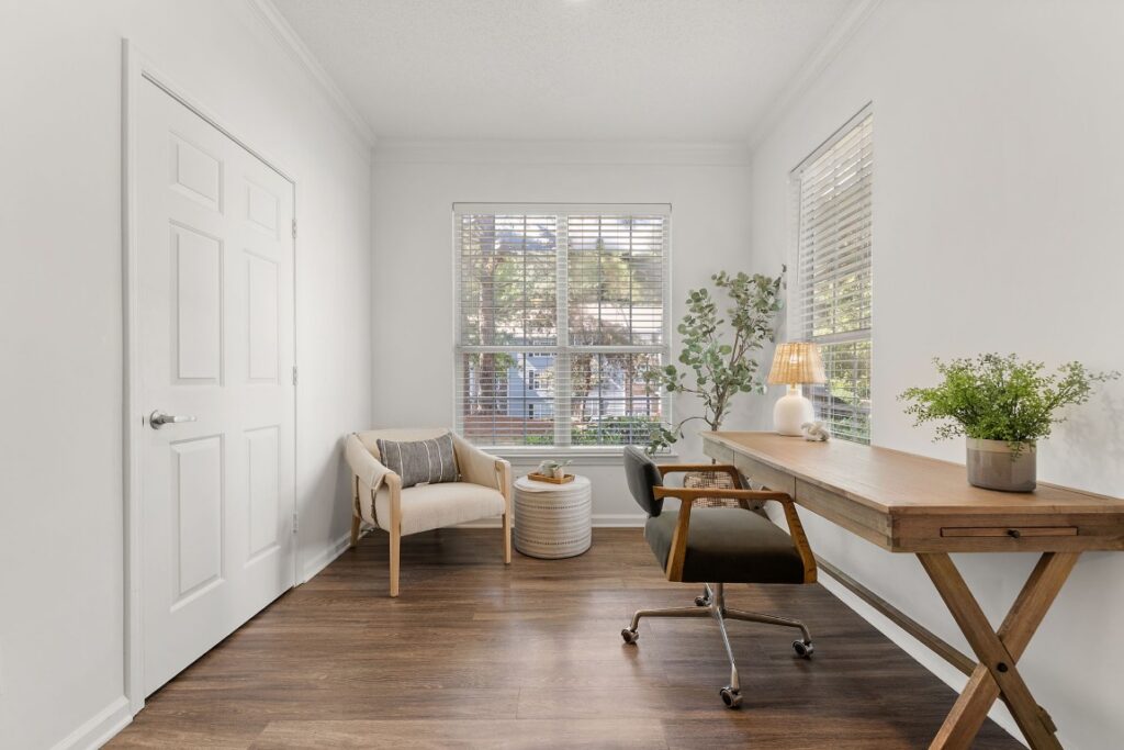 A bright home office with a wooden desk, rolling chair, and a cream-colored armchair in the corner.
