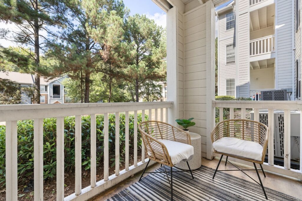 Two wicker chairs with white cushions on a striped rug on an apartment balcony overlooking trees.